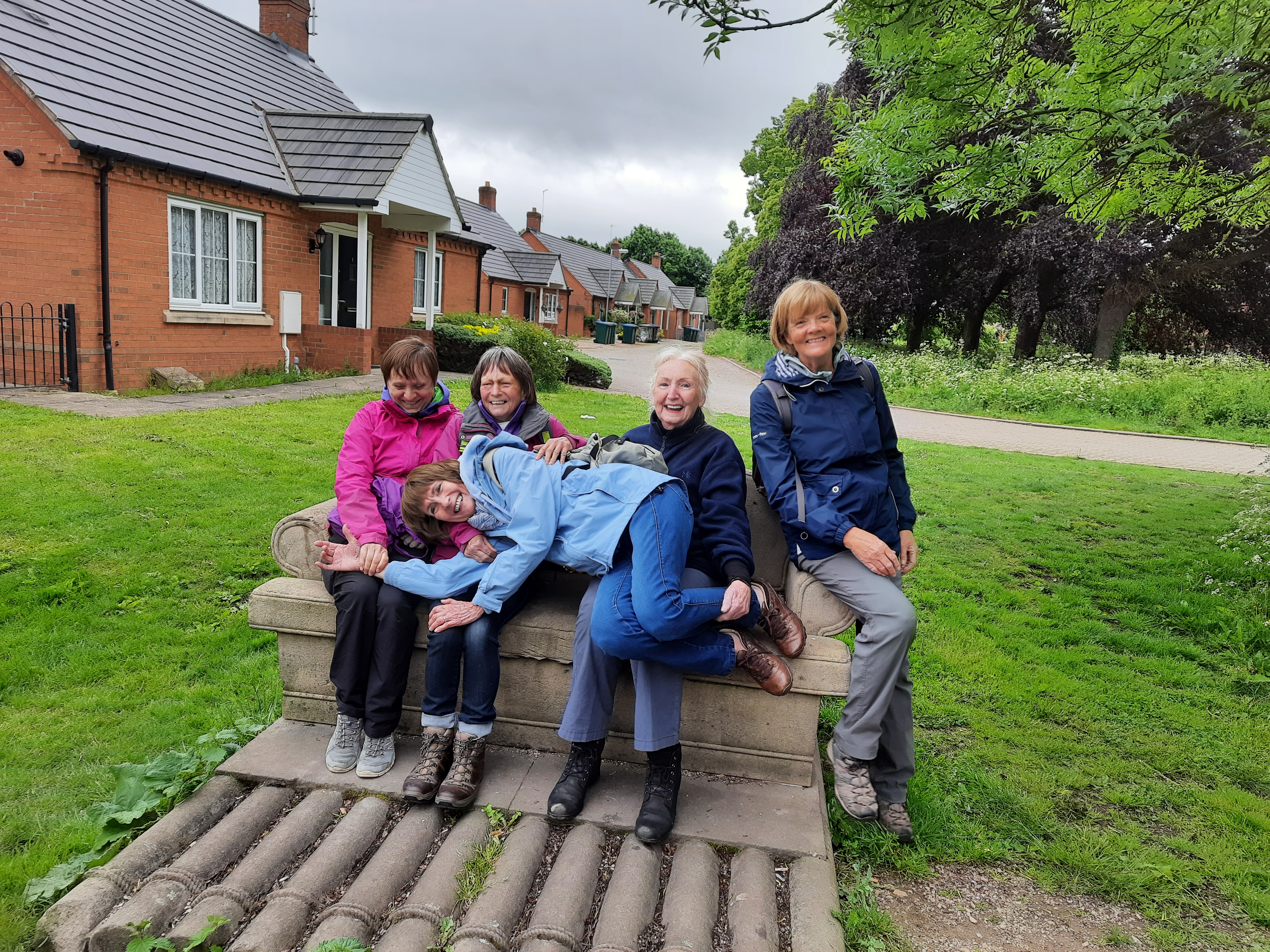 4 women sit on a bench with another lying across their legs. All are laughing.