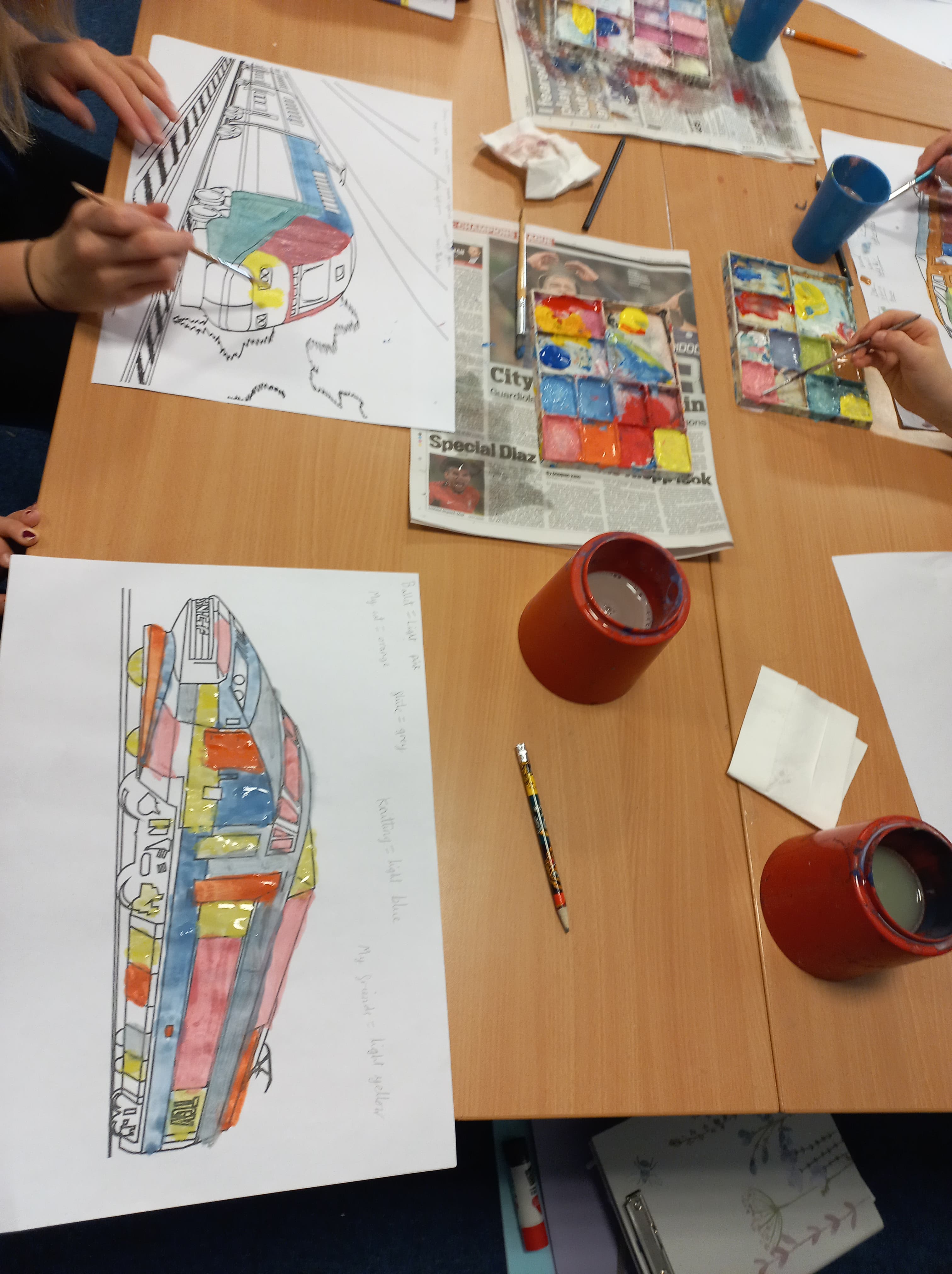 A table with a palette of paint with numerous colours in it. Hands painting trains in multicolours