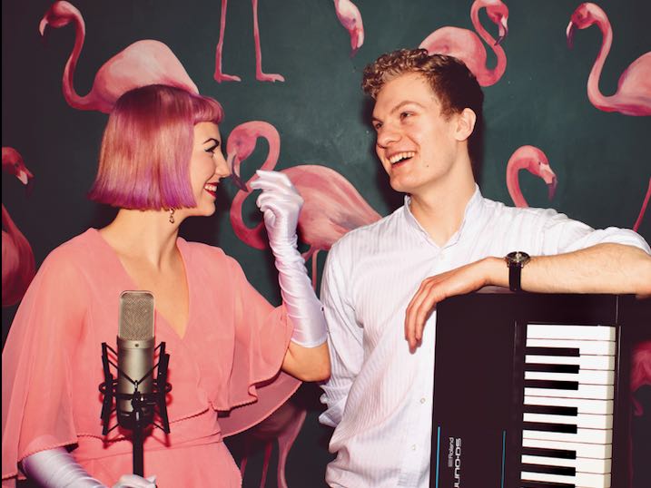 A woman in a pink dress with a microphone in front of her turns to a man leaning on a keyboard.  Both are smiling.