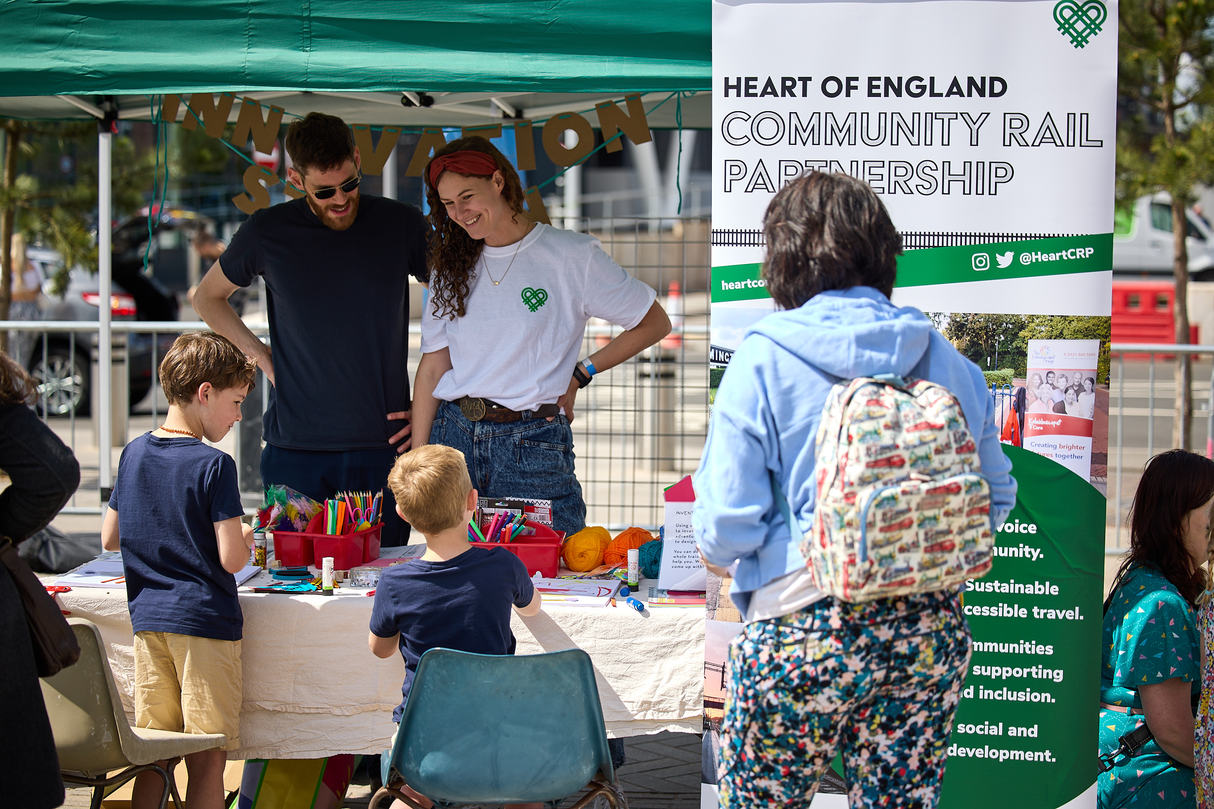 A man and a woman stand behind a table with art supplies, talking to two young boys about inventing their own trains.