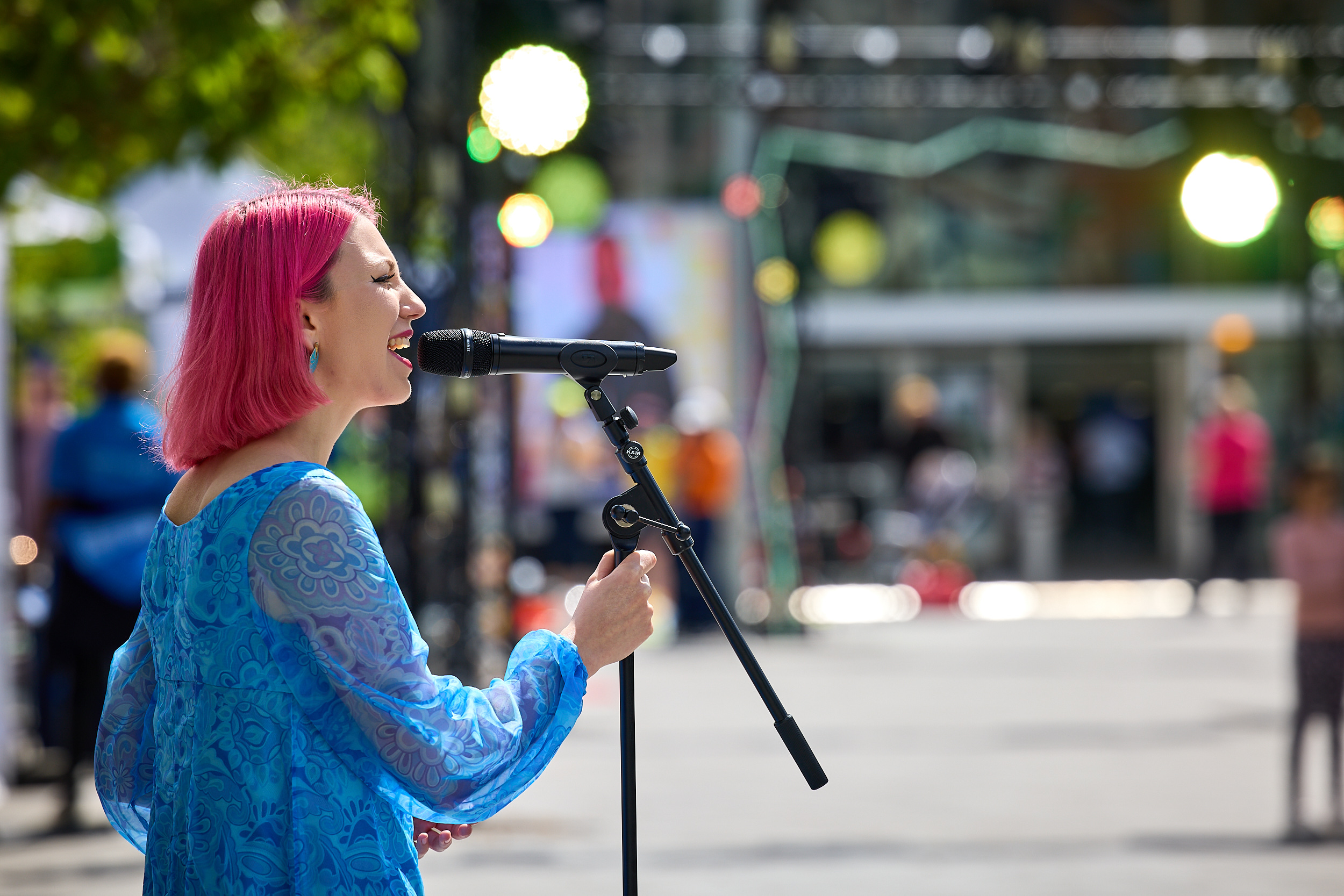 A lady with pink hair and a blue dress sings into a microphone in front of a blurred Coventry Stations