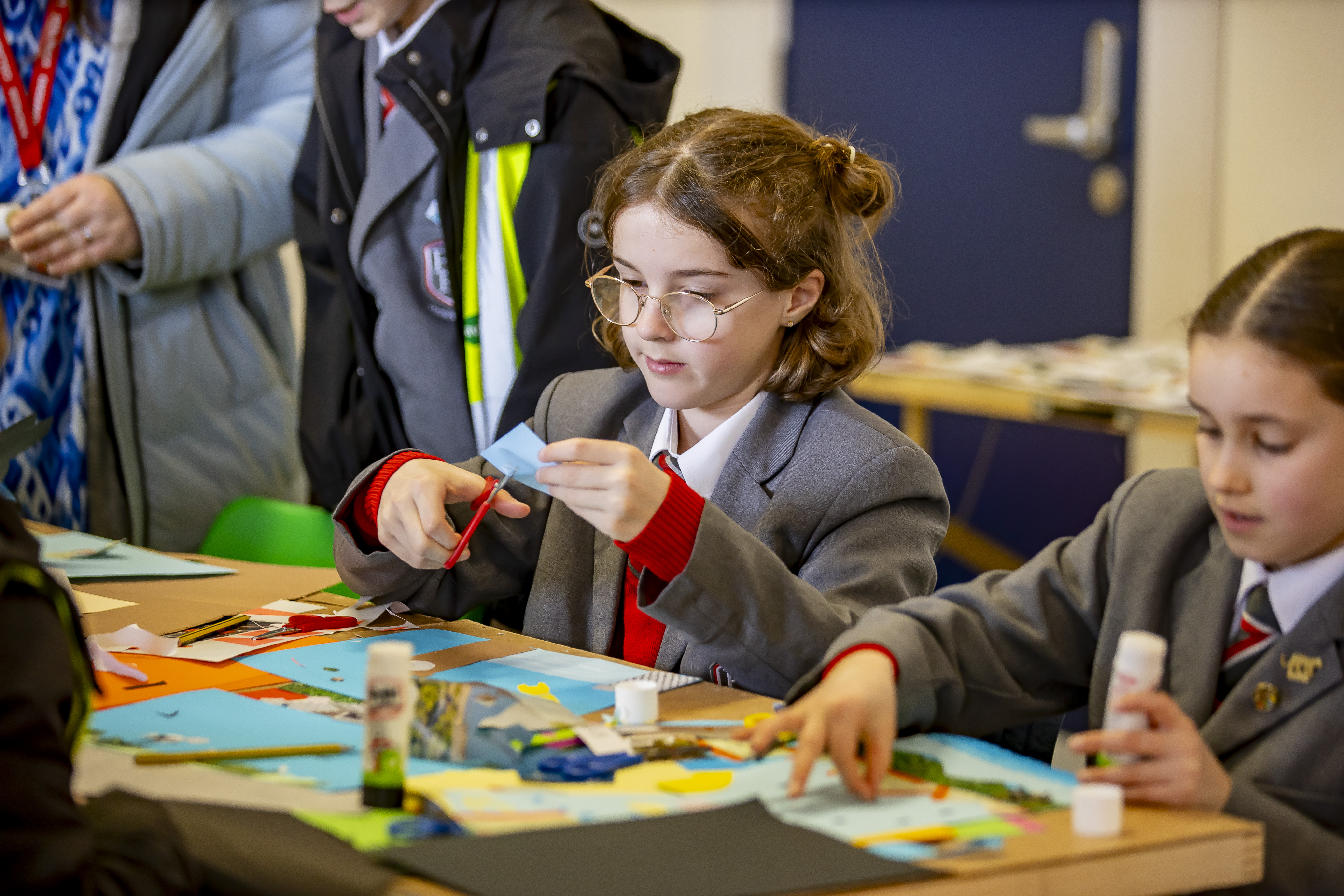 A school girl in a grey blazer cuts some paper for collage while he classmates sticks pictures to paper