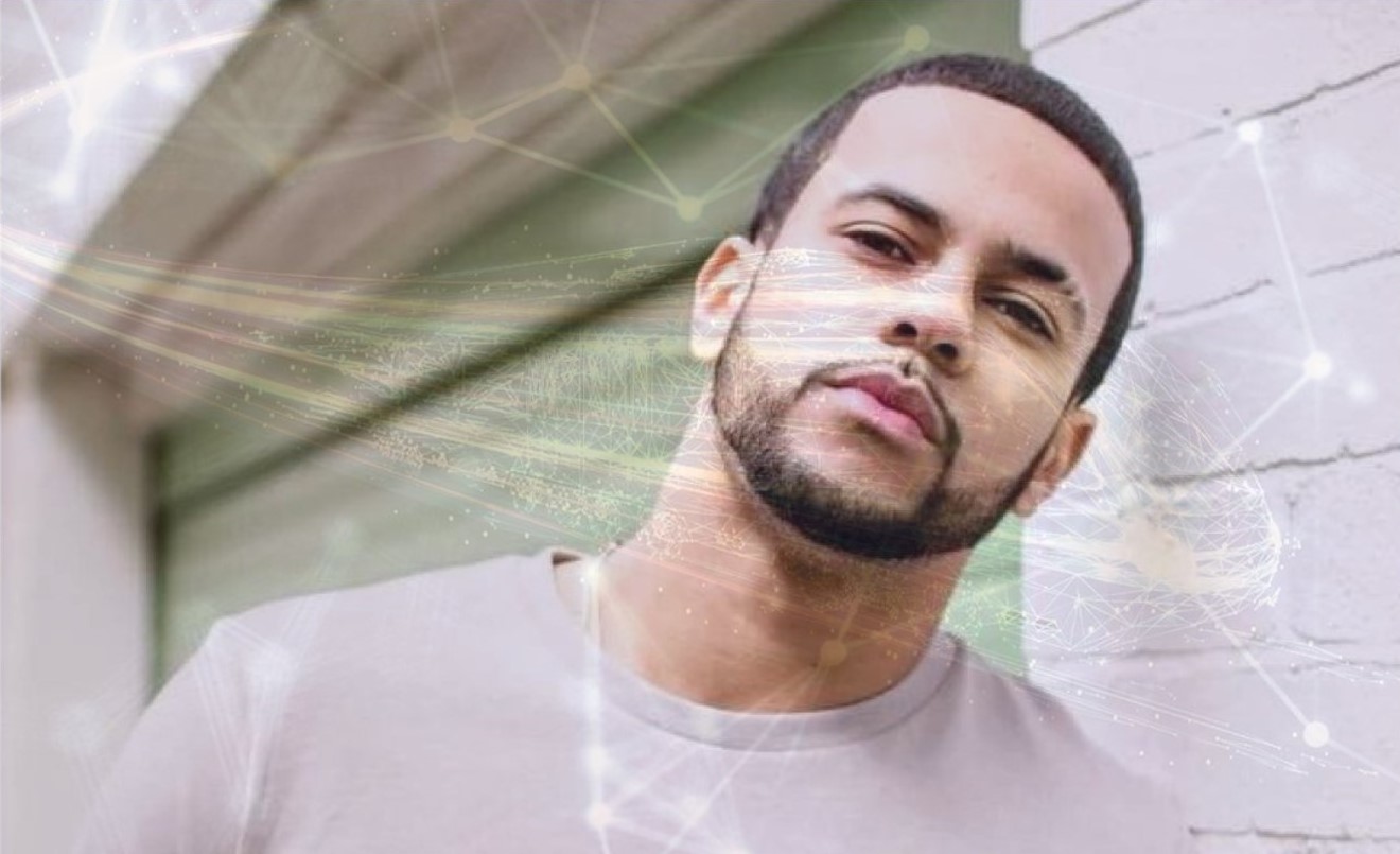 Picture of a young man with a goatee and a whit t-shirt, leaning towards a whitewall